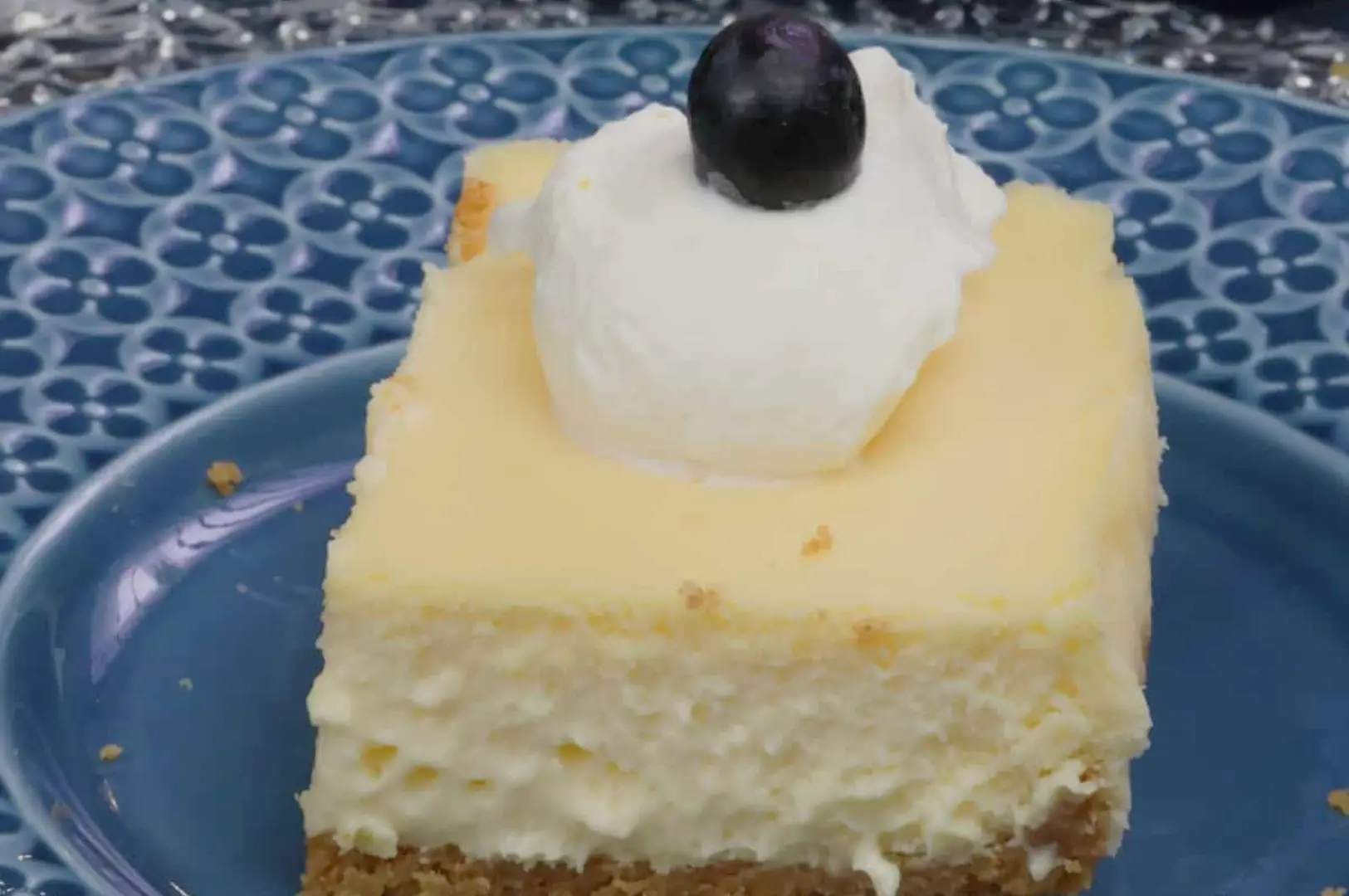 A piece of cheesecake topped with whipped cream and a blueberry, served on a blue plate with a decorative pattern.