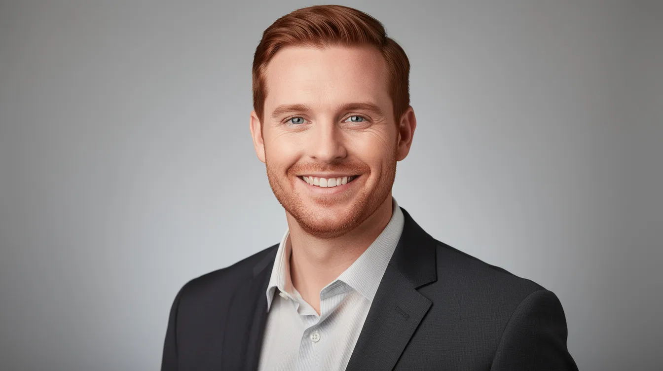 A professional headshot of a male real estate agent in his early to mid-30s features brownish-red short hair and light scruff, dressed in a dark tailored suit and crisp shirt, with an approachable smile against a neutral studio background. This image conveys a sense of confidence and professionalism, ideal for someone involved in the vibrant community of New Haven and its neighborhoods.