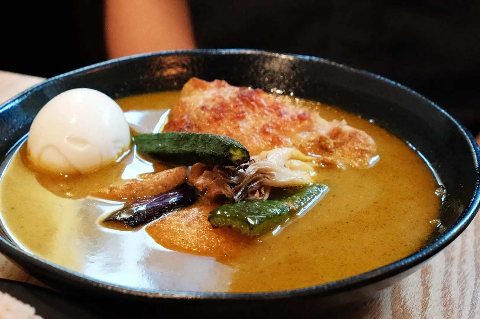 A black bowl of golden curry with a boiled egg, browned flatbread, eggplant, green vegetables, and mushrooms; a warm, appetizing meal.