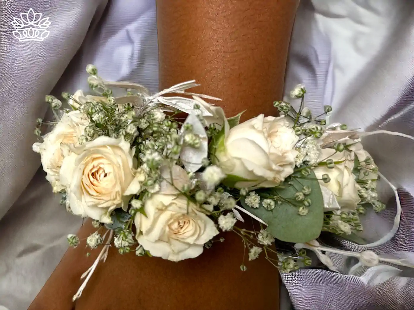 Elegant wrist corsage featuring creamy white roses interspersed with delicate baby's breath and soft green foliage, all tied with a thin white ribbon, gracefully worn against a silky silver fabric. Fabulous Flowers and Gifts - Matric Dance. Delivered with Heart.
