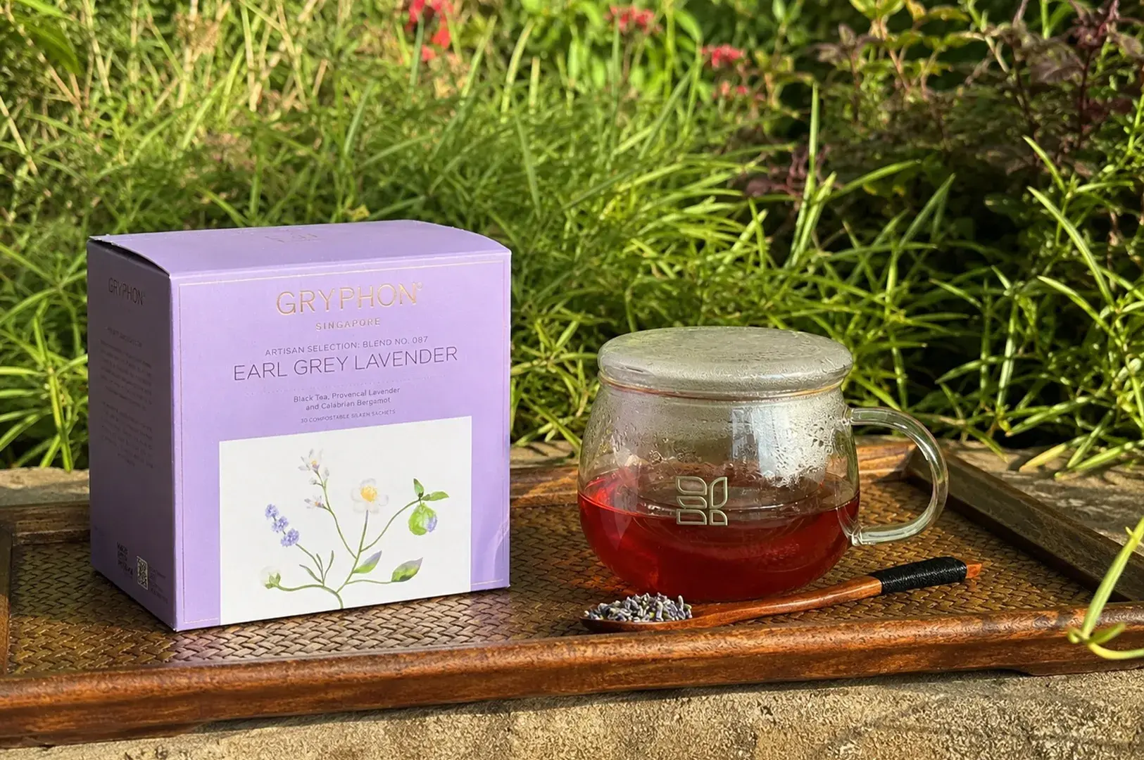 A lavender-colored cube box of Gryphon "Earl Grey Lavender" tea is displayed on a dark-toned woven bamboo tray outdoors against a background of soft green garden shrubs. Beside the box, a clear, rounded glass teacup with a matching glass lid contains a dark, amber-red brewed tea, showing subtle condensation near the rim. A small, elegant wooden spoon rests on the tray in front of the cup, holding a neat pile of dried purple lavender buds to highlight the aromatic ingredients of the blend.