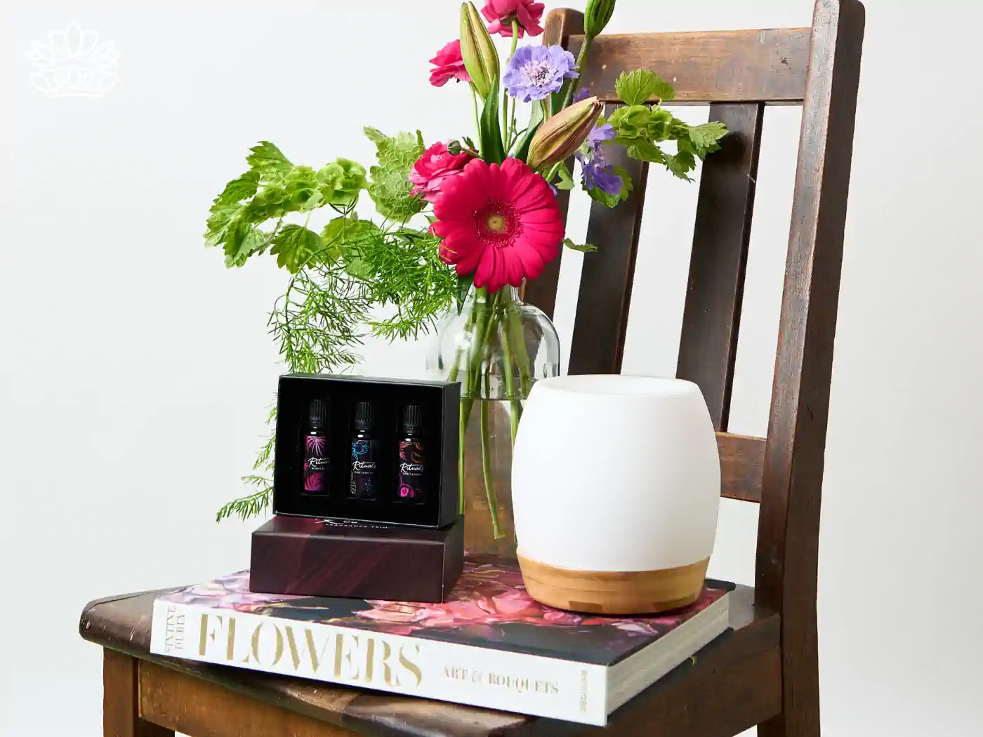 A tasteful housewarming gift setup displayed on an antique wooden chair, featuring a vibrant bouquet of fresh flowers in a clear vase, a box of essential oils, and a stylish aroma diffuser on top of a 'Flowers: Art & Bouquets' book. Perfectly blending nature with home comfort. Fabulous Flowers and Gifts.