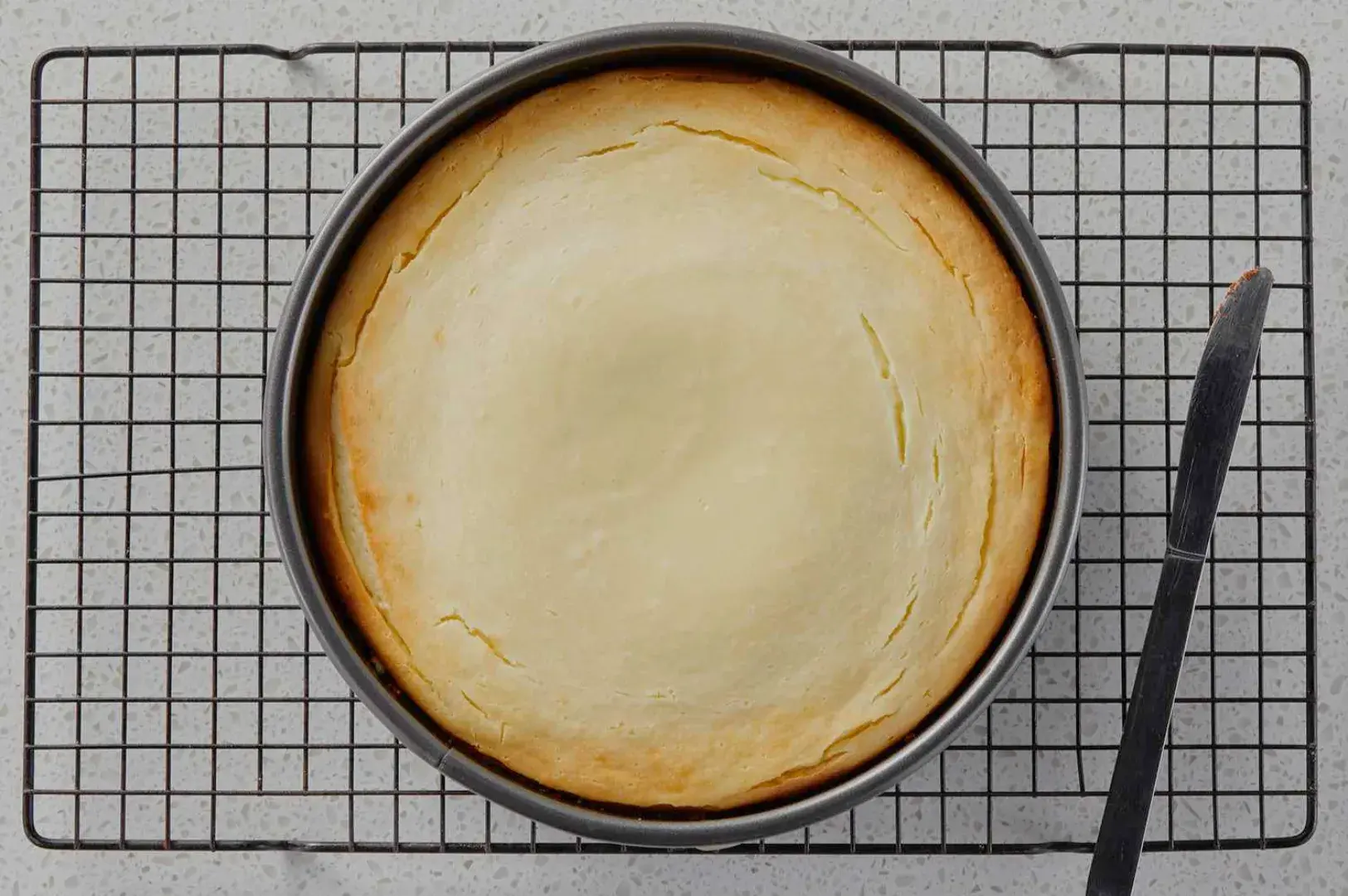 A freshly baked cheesecake cooling on a wire rack with a butter knife beside it.