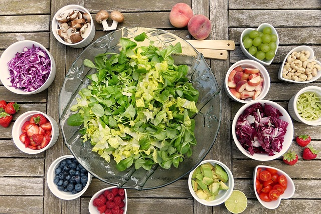 An image of a serving bowl of salad greens surrounded by small bowls of toppings, including blueberries, tomatoes, mushrooms, and green grapes.