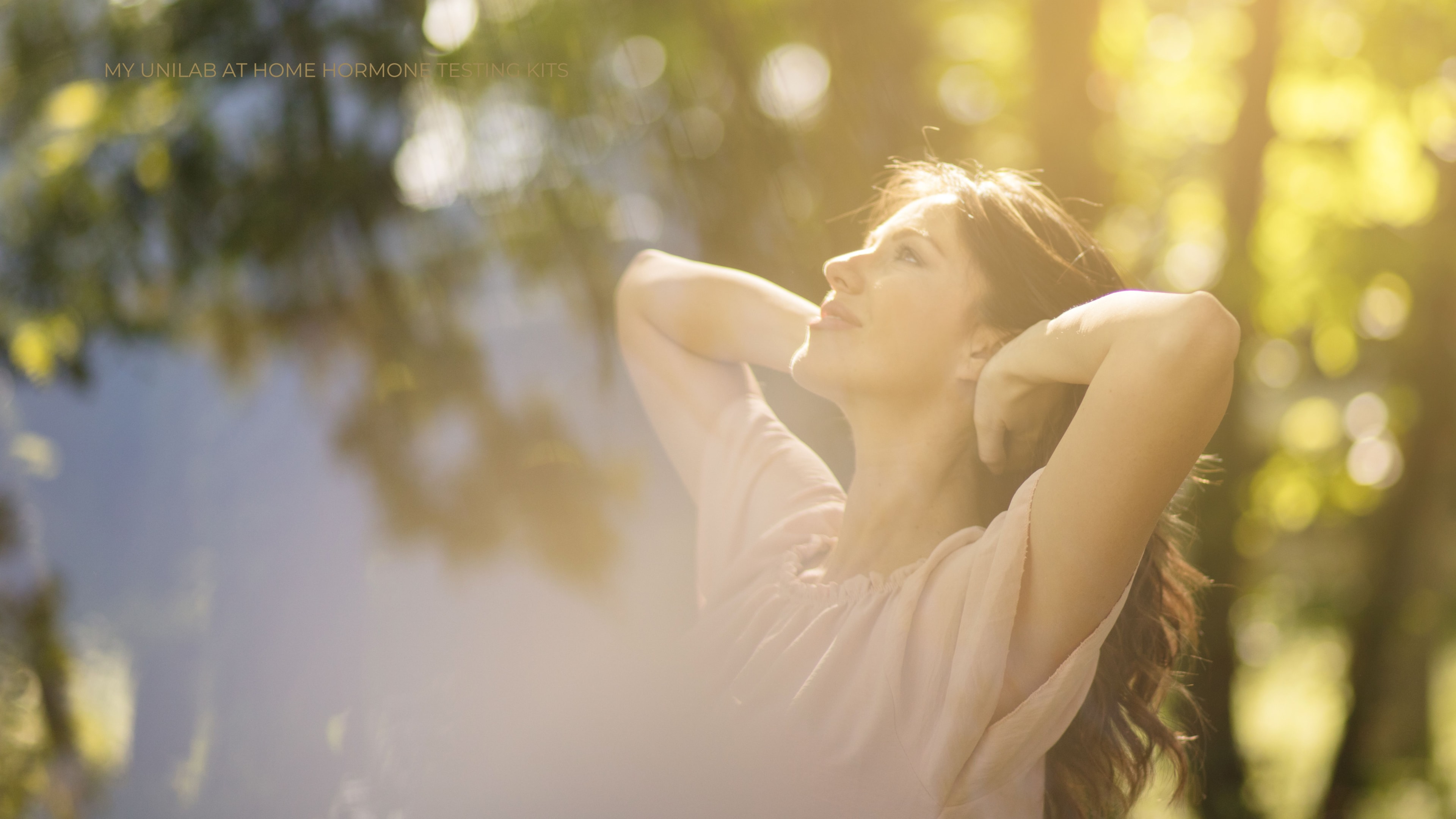 Female standing outside in nature with her arms lifted behind her head as she looks joyfully up towards the sun.