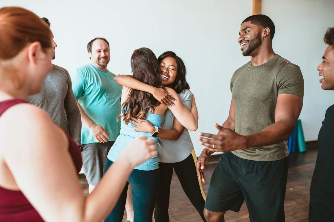 A group of people, with one person giving another a hug in the centre while others smile and watch. They are in a gym setting.