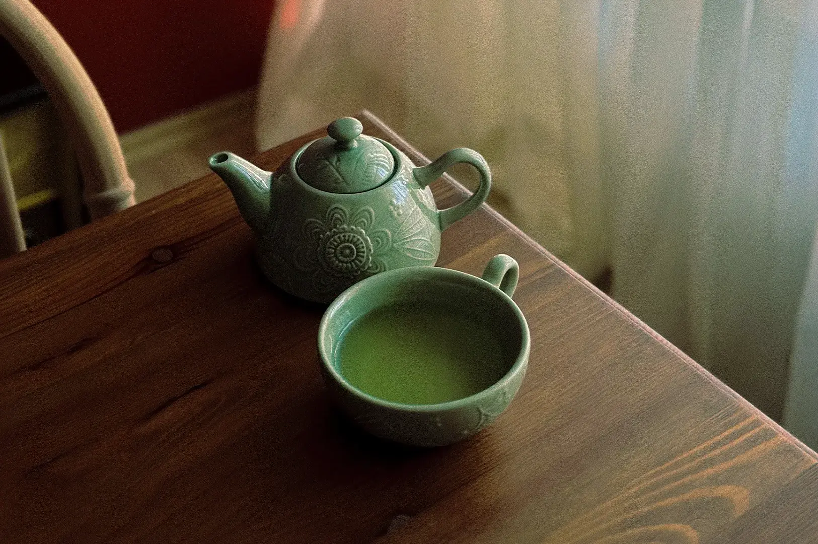 A cozy, close-up photograph captures a light green ceramic tea set resting on the corner of a dark, rustic wooden table. The set includes a small teapot decorated with intricate, embossed floral patterns and a matching wide-rimmed teacup filled to the brim with vibrant, opaque green tea. The scene is illuminated by soft, natural light streaming in from the right, partially filtered by a sheer white curtain in the background, which creates a warm, serene, and inviting atmosphere.