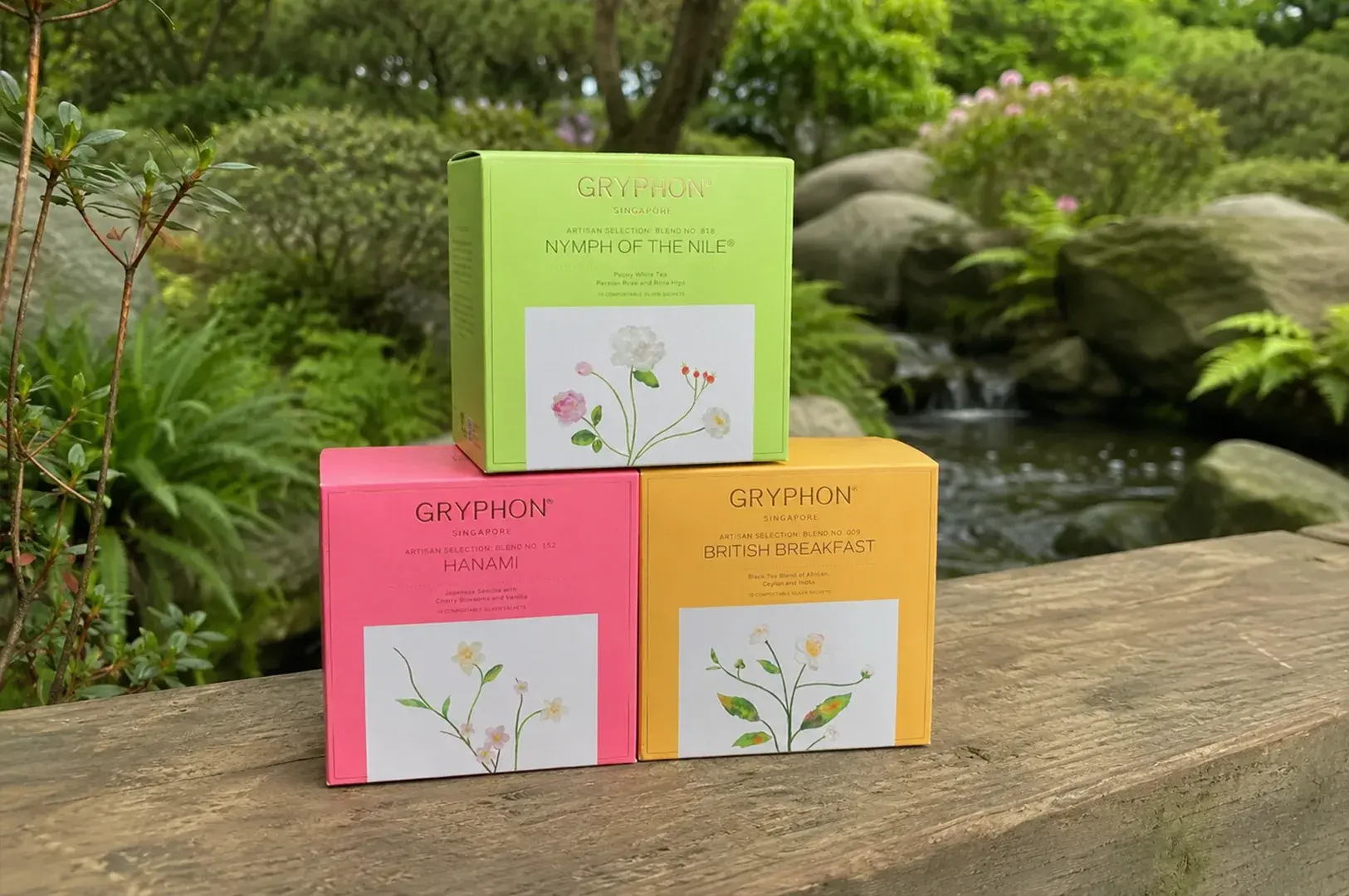 Three colorful, cube-shaped Gryphon tea boxes are stacked in a neat pyramid on a thick, rustic wooden railing with a traditional Japanese garden setting in the background. The top box is a bright lime green ("Nymph of the Nile"), resting on a hot pink box ("Hanami") on the left and a warm yellow box ("British Breakfast") on the right. Each box features a distinct botanical watercolor illustration on its front panel, set against a serene background of a stone-lined pond, lush greenery, and a small, cascading waterfall.