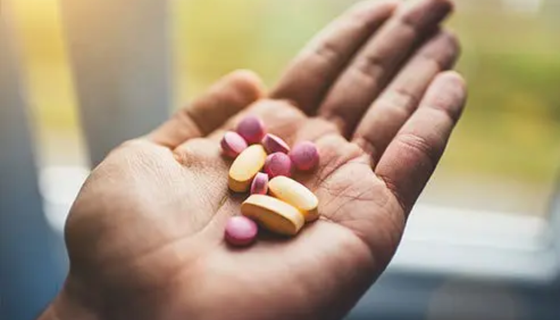En åpen hånd som holder rosa og gule kosttilskuddstabletter i naturlig dagslys nær et vindu.