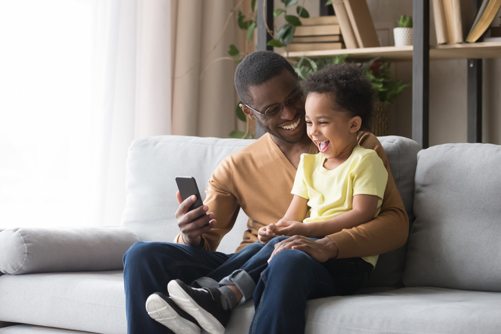Cheerful young dad sitting on the sofa with his daughter in his lap looking at a cell phone.