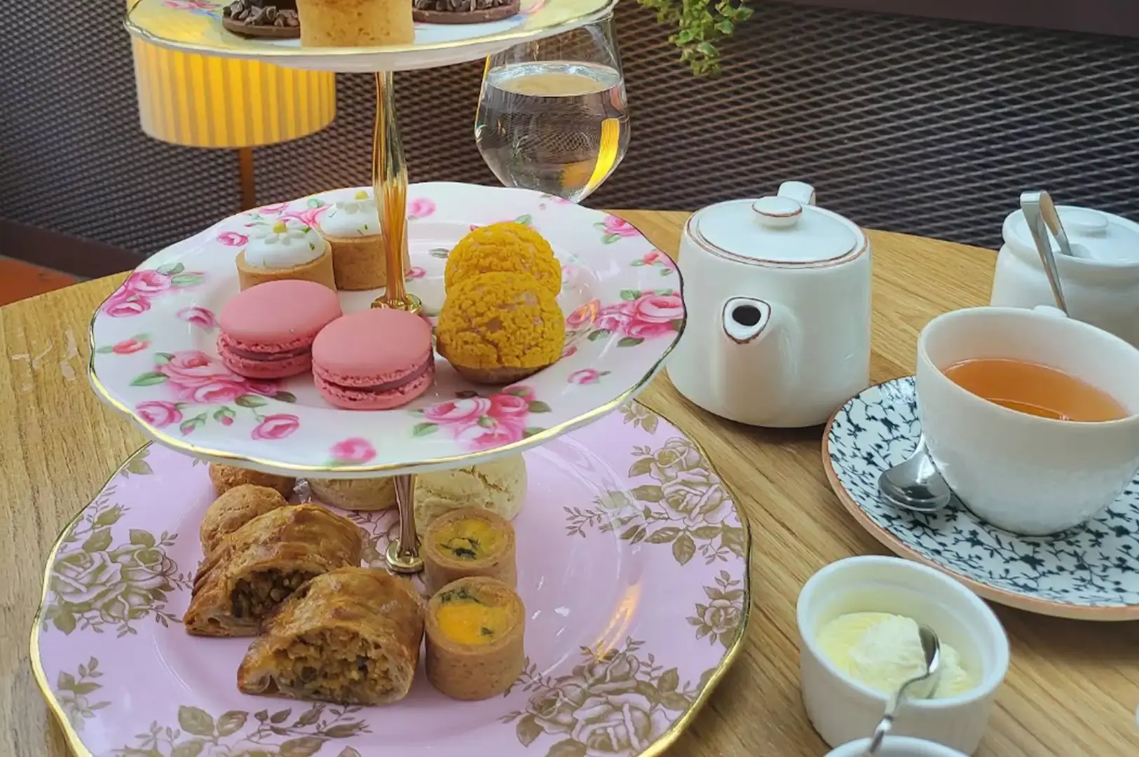 A multi-tiered floral afternoon tea stand filled with pink macarons, golden choux pastries, savory tarts, and sliced rolls, served on a light wooden table alongside a white teapot, a cup of amber tea, and small dishes of clotted cream.