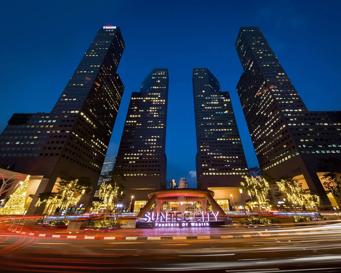 Night view of Suntec City, with five illuminated skyscrapers forming a semicircle. The foreground features the Fountain of Wealth with glowing lights.