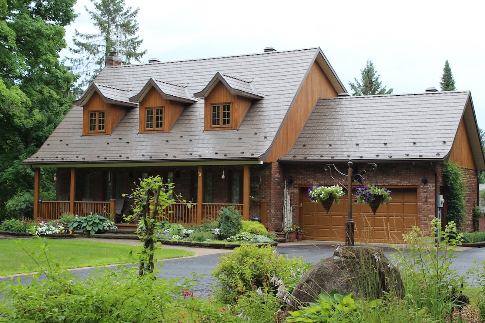 A picture of a home with a steel roof in a dark brown which matches the colour of the home. The house is surrounded by trees and a well-maintained front yard with lots of flowers and plants. 