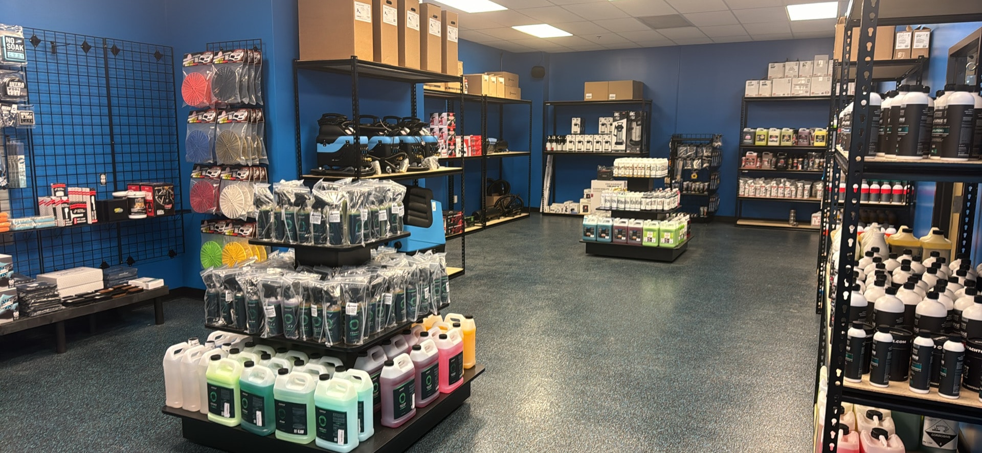 A car enthusiast is intently watching a tutorial video on his smartphone while examining various detailing products in a car care store. The shelves are stocked with premium car cleaning products, including ceramic coatings and polishing compounds, designed to enhance and protect vehicles.