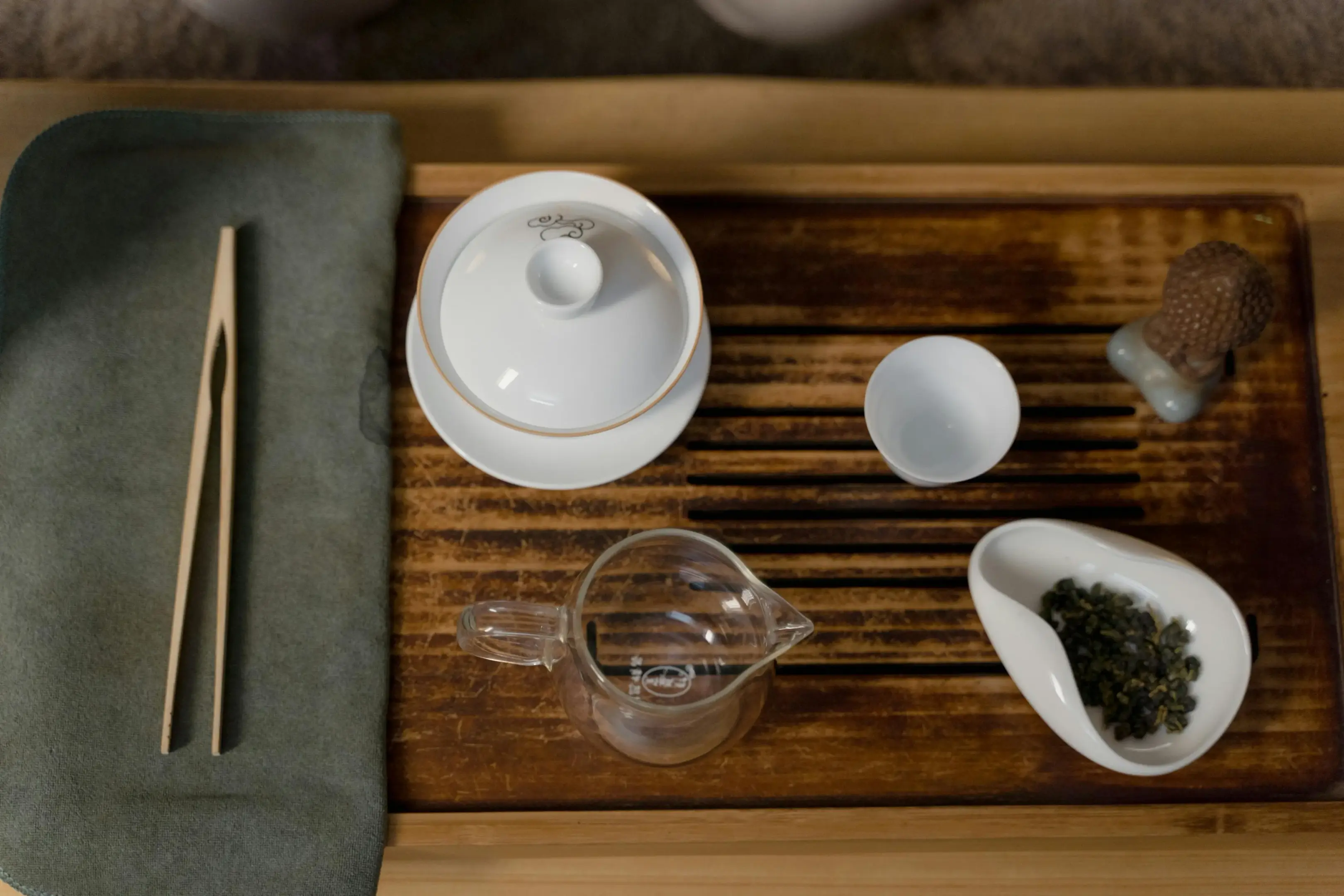 This image showcases a traditional Gongfu tea set arranged neatly on a wooden slatted tea tray. The collection includes a white lidded gaiwan, a small matching teacup, a glass fairness pitcher, and a ceramic scoop holding loose-leaf tea.