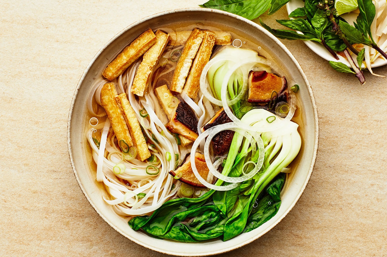 Top-down view of a bowl of Vietnamese vegetarian pho with tofu, rice noodles, bok choy, and onion slices on a light-colored surface.