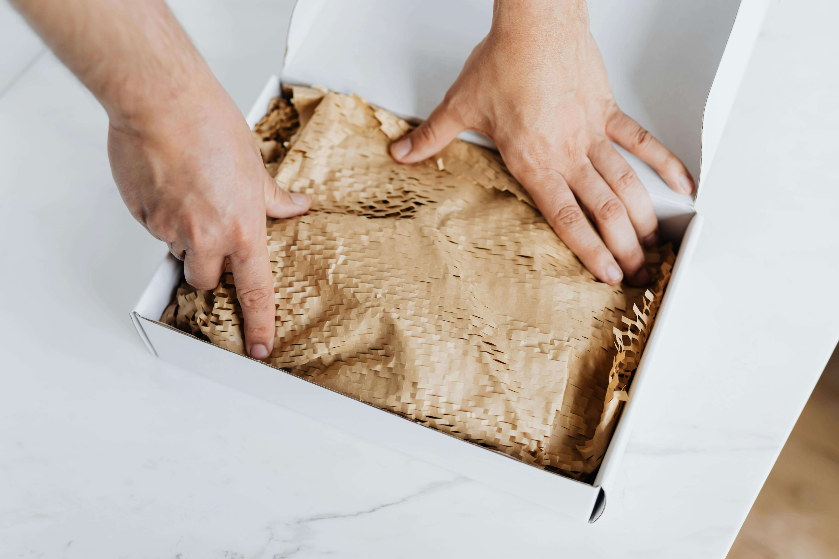 Hands arranging protective wood shavings in a white gift box on a marble countertop.