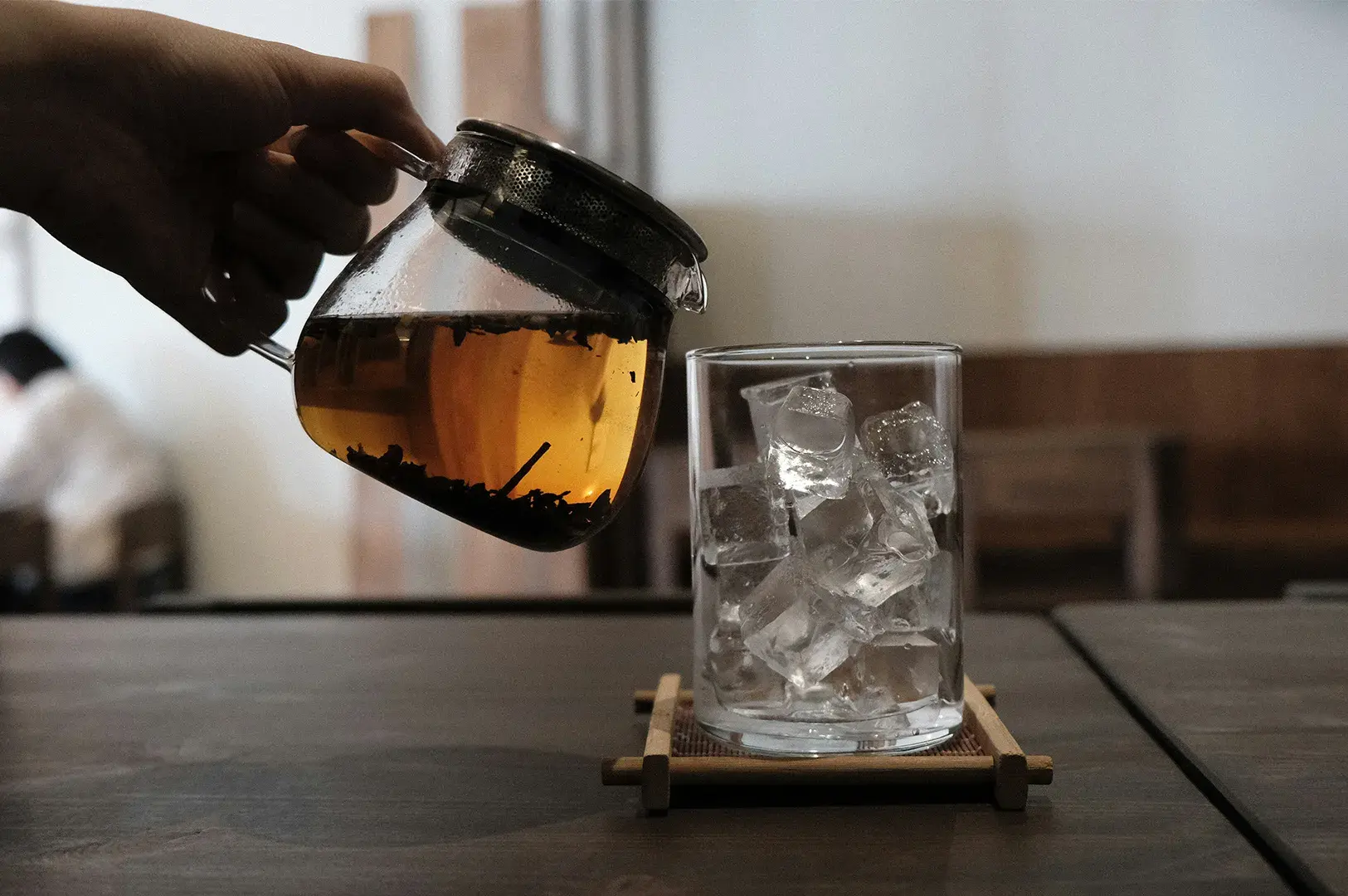 Glass teapot pouring brewed tea into a glass of ice on a bamboo coaster.