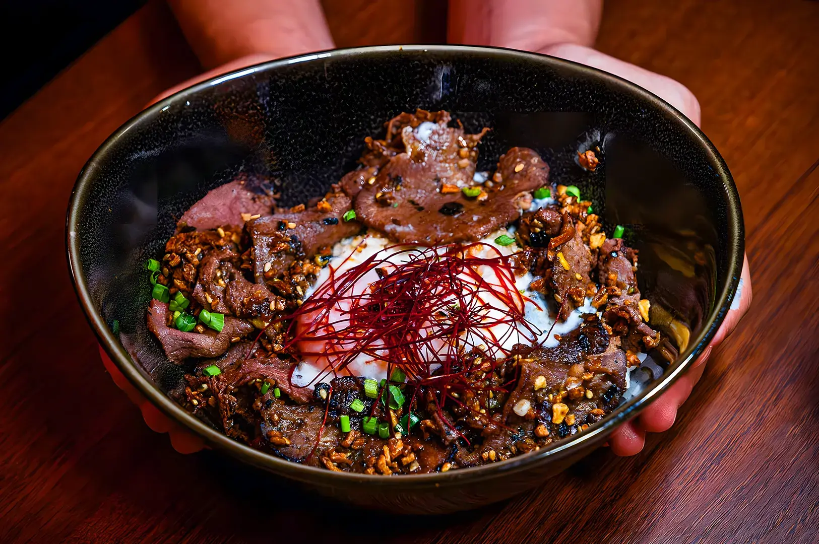 A bowl of savory beef donburi topped with a poached egg, garnished with green onions, red chili threads, and crispy garlic, held in hands on a wooden table.