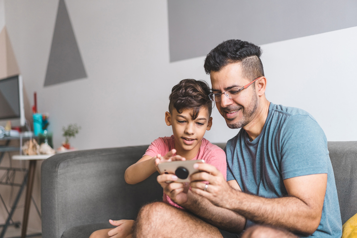 Cute little boy and his dad sitting on a sofa looking at a cell phone.  