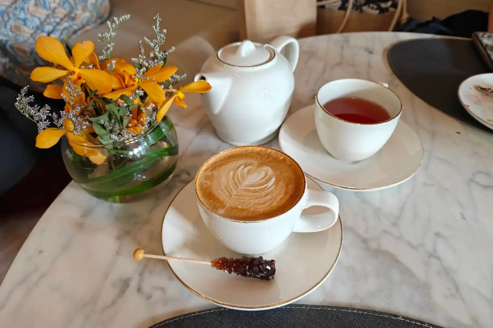 A white marble tabletop is set with a white ceramic teapot, a teacup filled with amber-colored tea, and a white coffee cup filled with latte art accompanied by a rock candy stir stick. A small glass vase containing bright yellow orchids and delicate filler flowers adds a touch of color near the beverages.