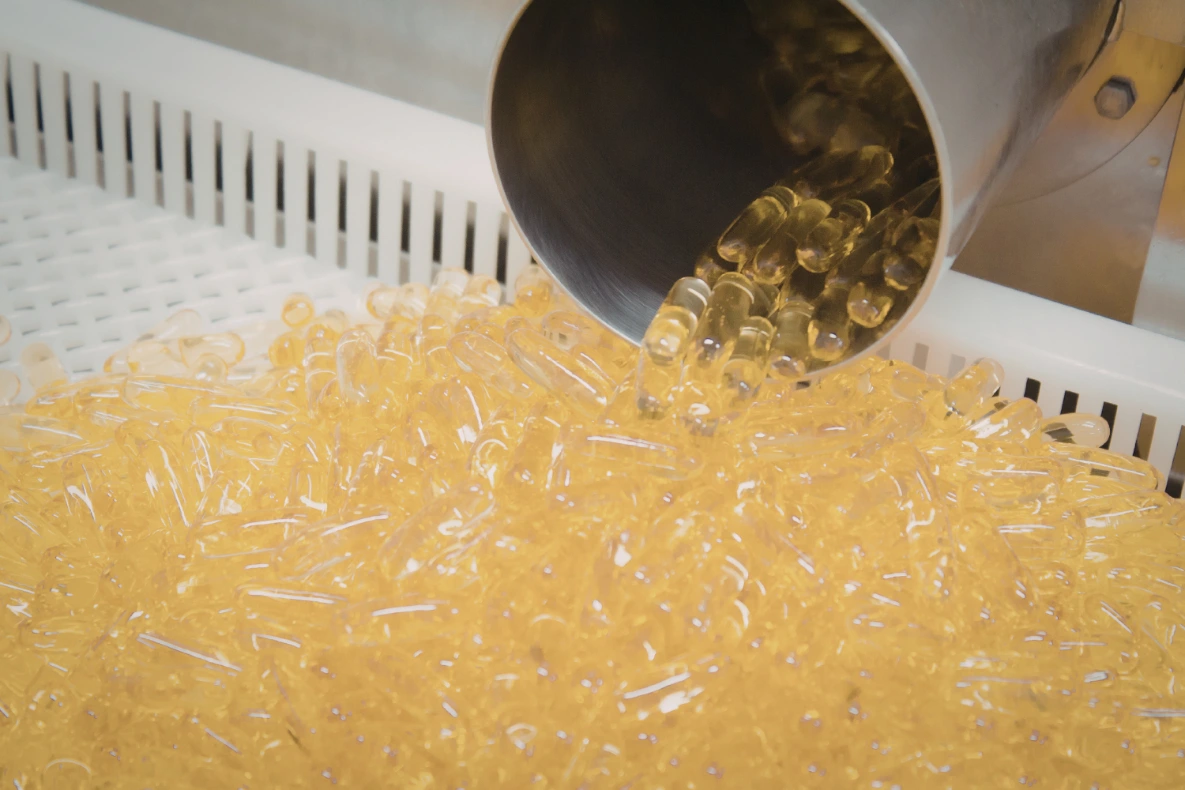 A metal cylinder pours a large quantity of yellow gel capsules onto a white, slotted surface.