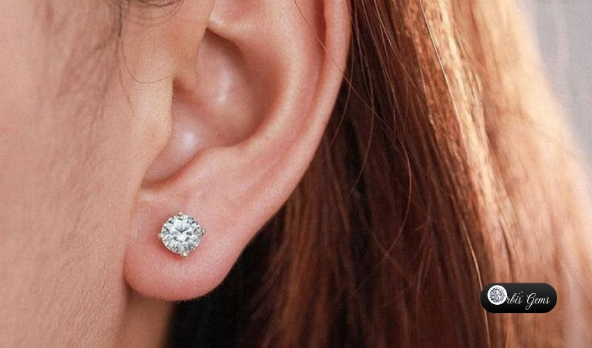 Close-up of a woman's ear wearing a sparkling moissanite solitaire earring with a silver setting, by Orbis Gems in a post about rise of moissanite stud earrings