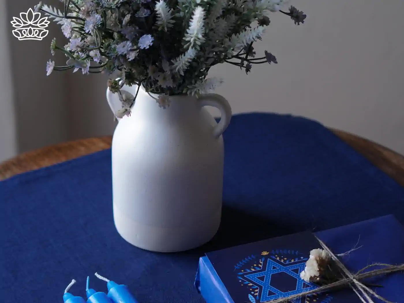 A white ceramic vase filled with delicate purple and white flowers, sitting on a blue cloth alongside Hanukkah candles and a wrapped gift. Fabulous Flowers and Gifts - Hanukkah Flowers.