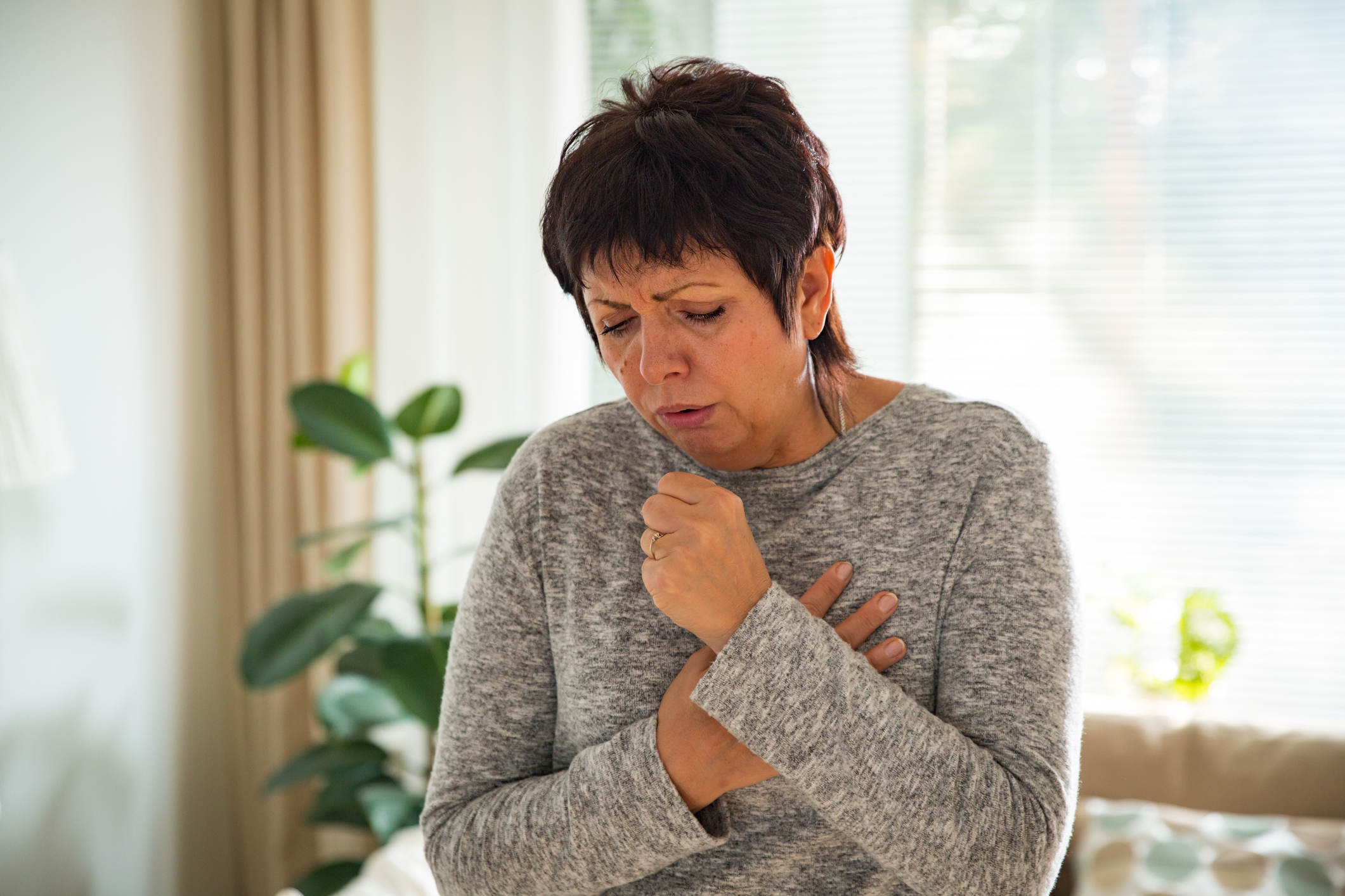 An image of a woman with a dry cough holding one hand over her chest and the other hand or fist to her mouth. 