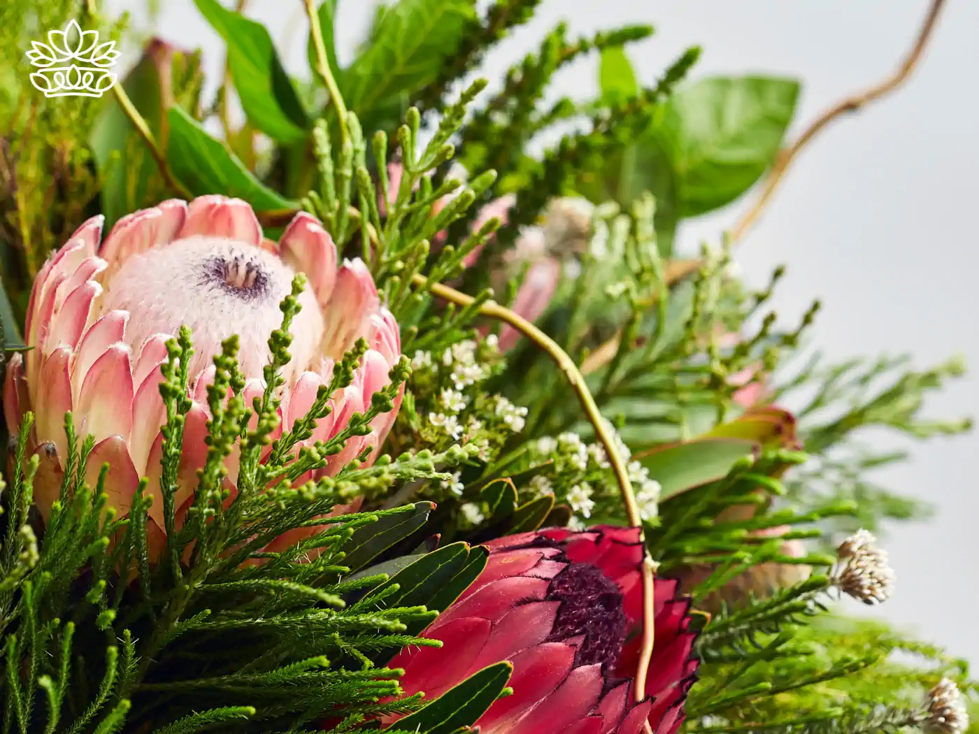 A close-up of a stunning flower arrangement featuring pink protea flowers surrounded by vibrant greenery and delicate white blossoms. This lush display showcases the unique beauty and texture of proteas, making it a standout piece in any floral collection. All Flower Arrangements Collection. Fabulous Flowers and Gifts.