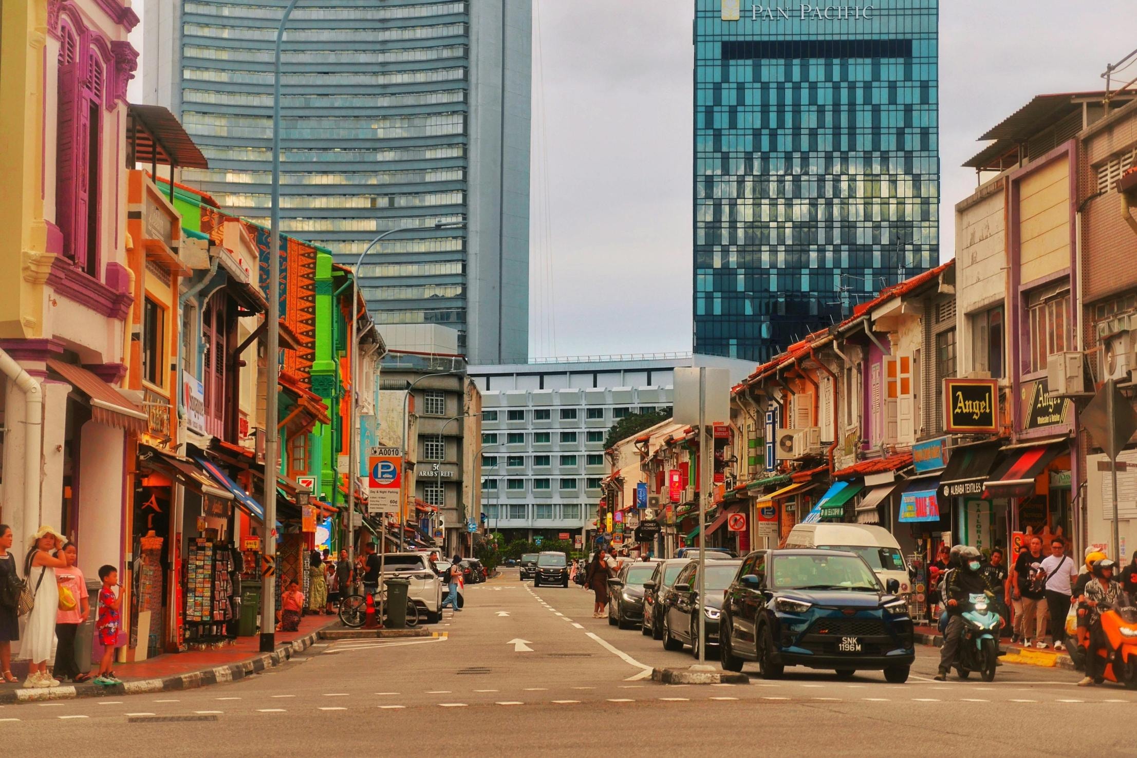 A busy street is flanked by rows of vibrant, multi-colored shophouses, with pedestrians walking along the sidewalks and cars navigating the road. Towering modern skyscrapers, including the prominent Pan Pacific building, rise in the background, creating a striking contrast with the historic low-rise architecture below.