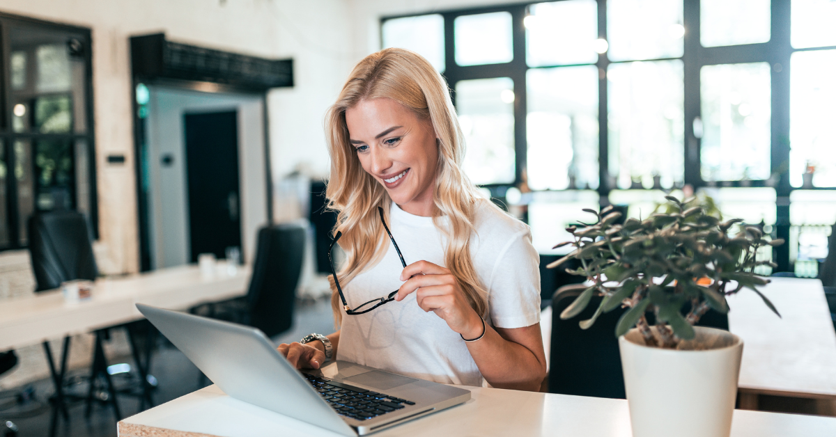 A smiling woman working on her laptop in a bright office; exploring a self-employed tax calculator.