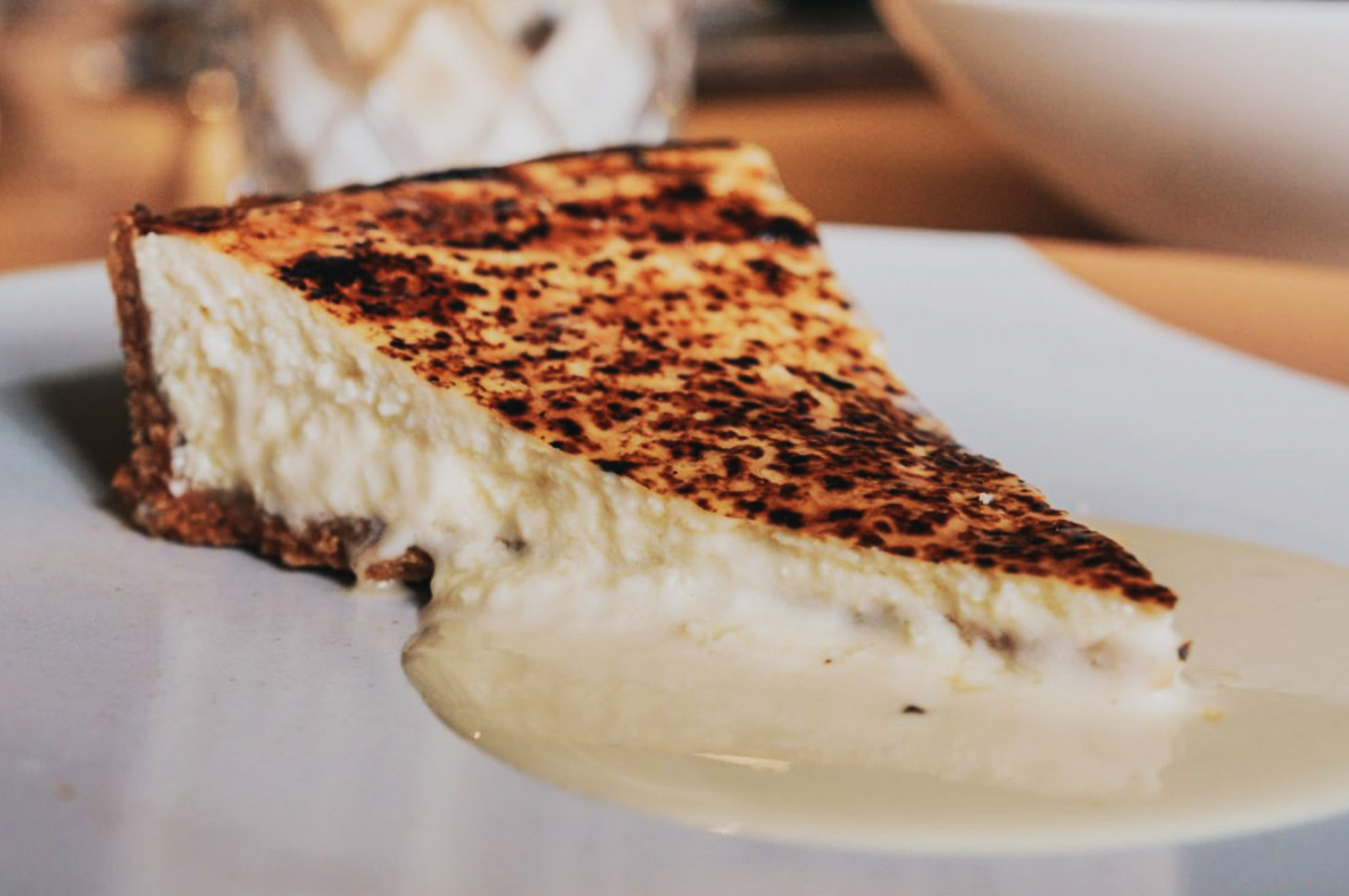 A slice of creamy cheesecake with a burnt caramelized top sits on a white plate, exuding a rich and indulgent texture.