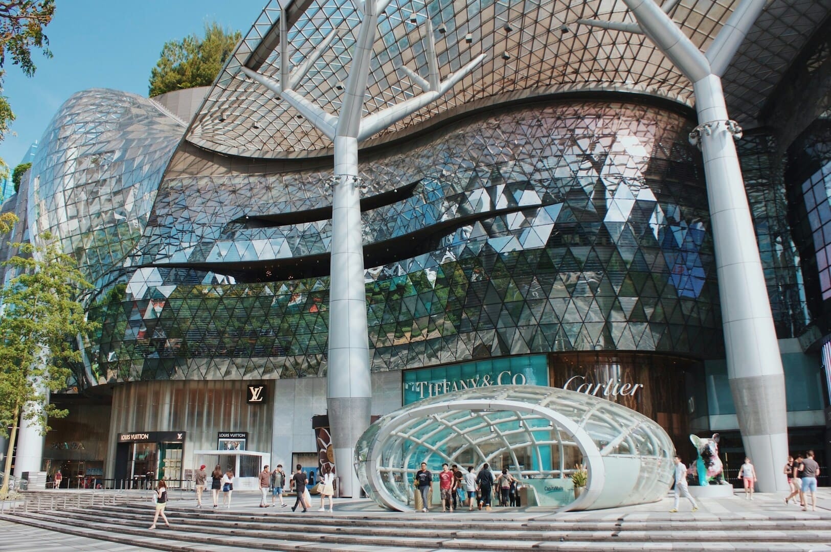 Futuristic mall entrance at Ion Orchard with reflective glass facade and intricate patterns. Elegant stores, people walking, and a curved glass canopy create a vibrant, modern scene.