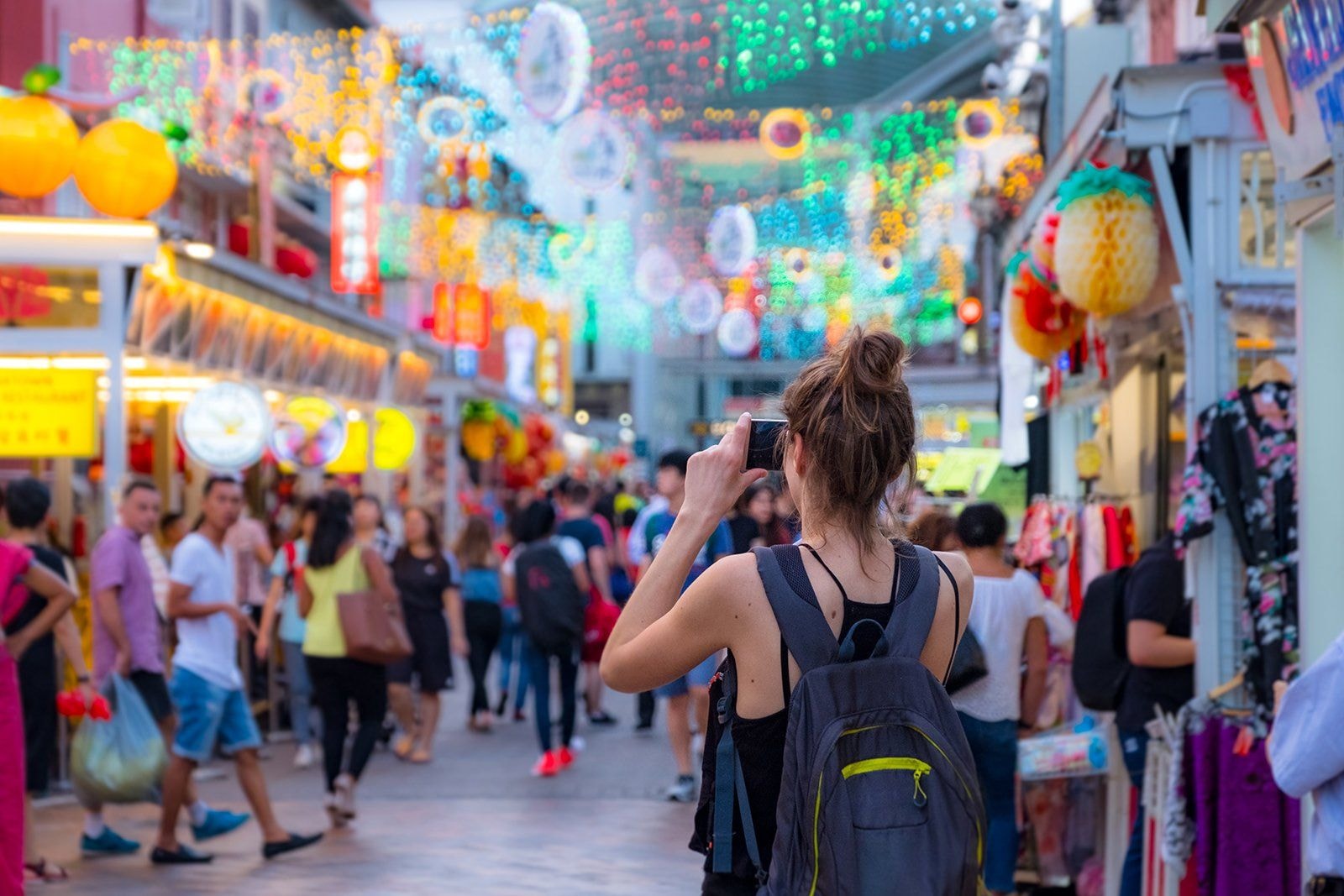 A woman with a backpack stands in a crowded pedestrian street, holding up a camera to capture the vibrant atmosphere. The scene is illuminated by glowing festive lights and colorful fruit-shaped lanterns hanging above various market stalls and shops.