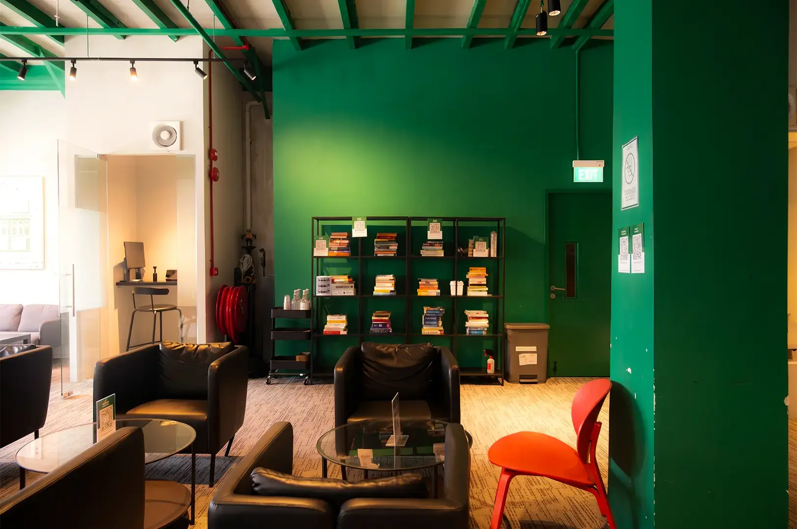 A view of Monk's Brew Club with black cushioned chairs and glass tables arranged inside a green and white colored walls and columns, bright sunlight illuminating the left side.