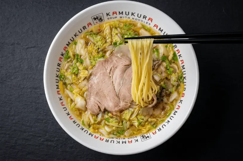 A hearty bowl of late-night ramen resting on a wooden counter, generously topped with thick chashu pork slices, a whole soft-boiled egg, bamboo shoots, chopped scallions, bonito flakes, and a large, crisp sheet of nori seaweed.
