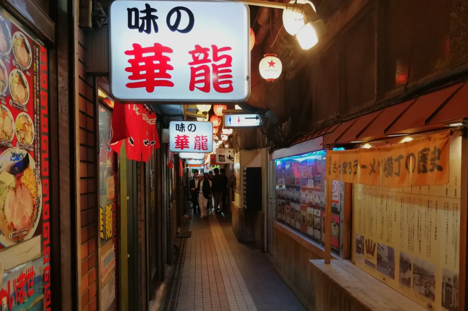 A narrow alleyway with neon signs and illuminated lanterns, lined with shops and restaurants.