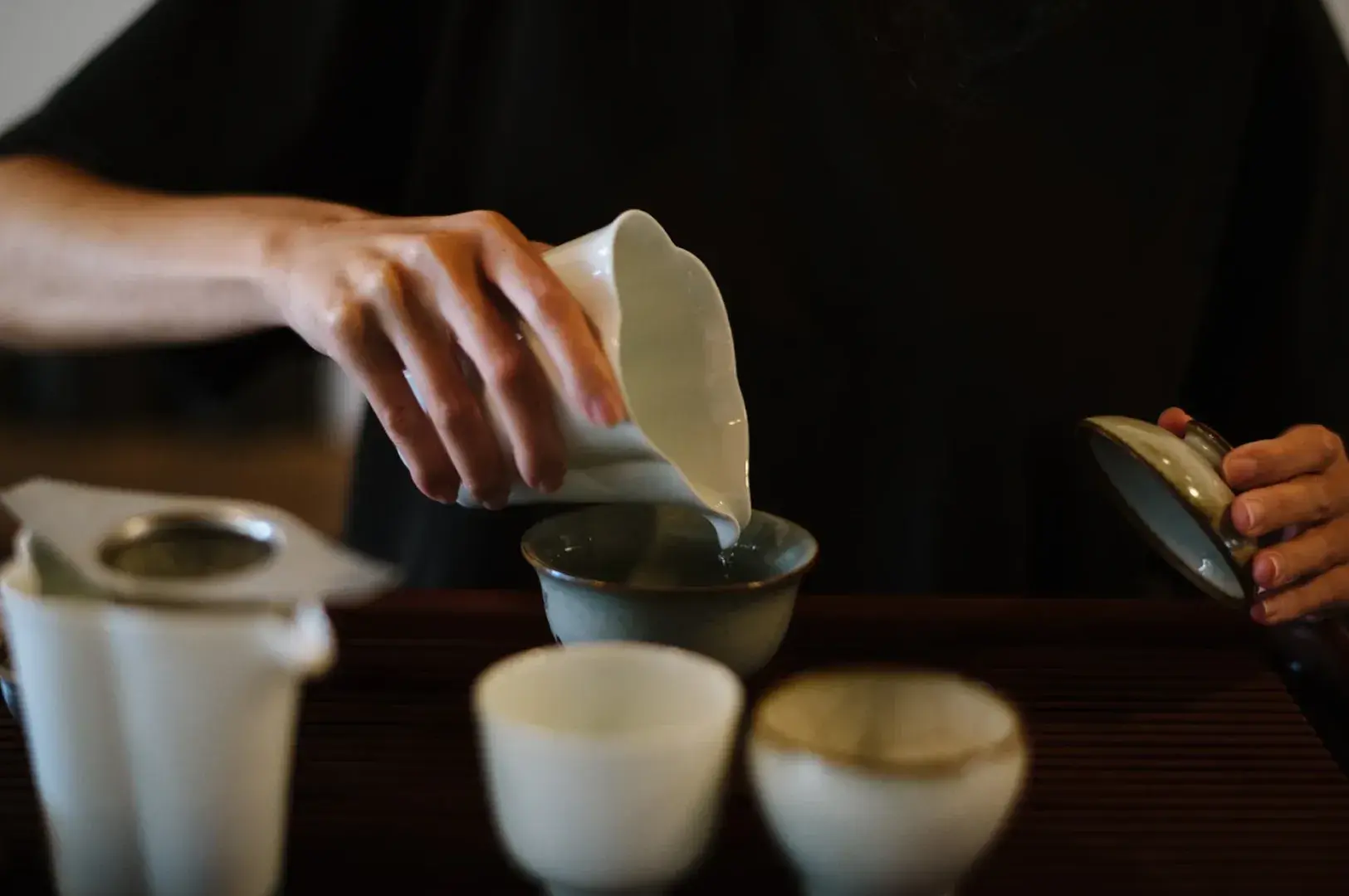 Close-up of hands pouring steeped tea from a ceramic pitcher into tasting cups.
