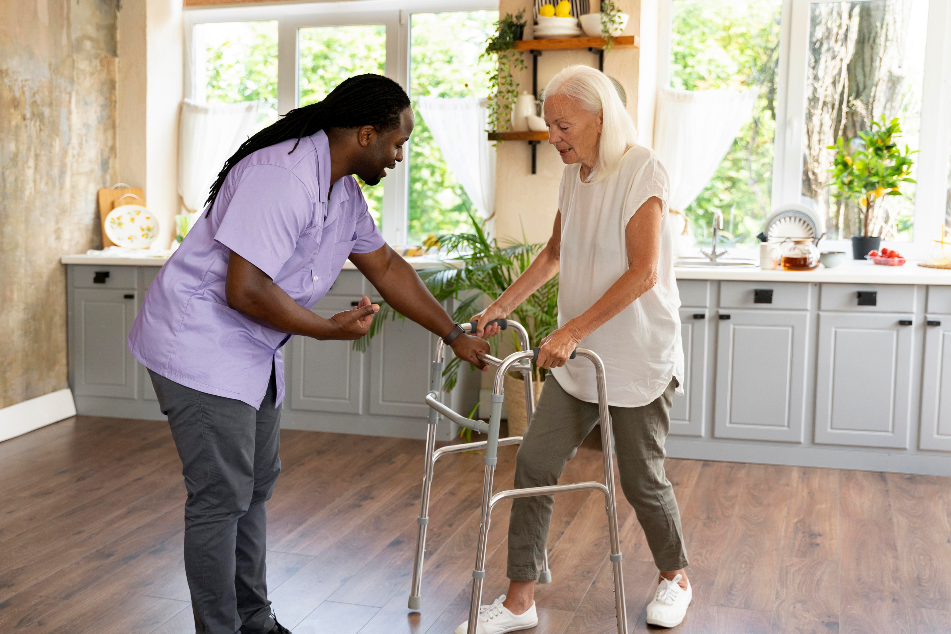 An older woman is in a kitchen, using a walking frame. A male healthcare worker supports her whilst she walks by holding the front of the walking frame. 