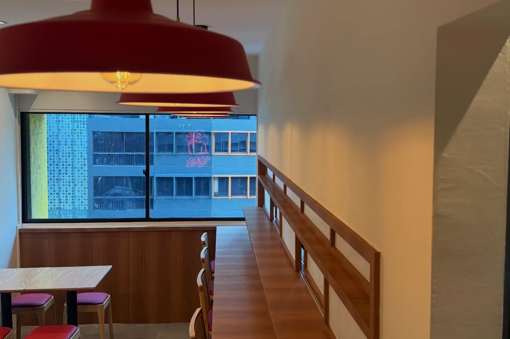 A modern cafe interior featuring wooden seating, red pendant lights, and a large window with a view of an urban building.