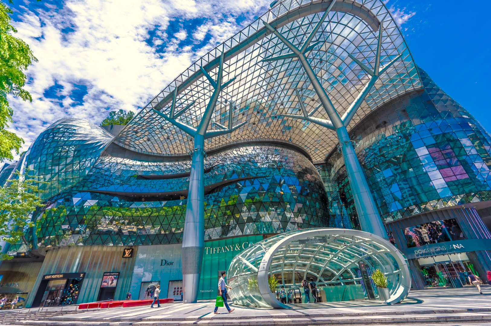 Futuristic glass facade of a shopping mall under a clear blue sky. Reflective geometric patterns and curved architecture create a vibrant, modern aesthetic.