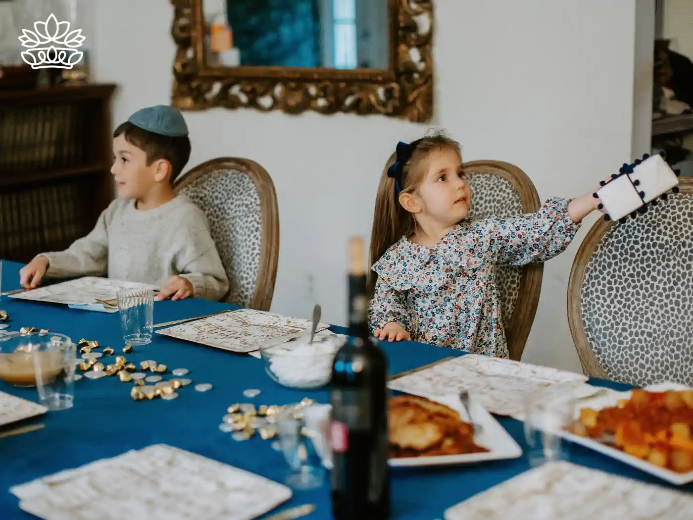 Two children sitting at a table, one passing a gift to the other, surrounded by Hanukkah decorations. Fabulous Flowers and Gifts - Hanukkah Collection.