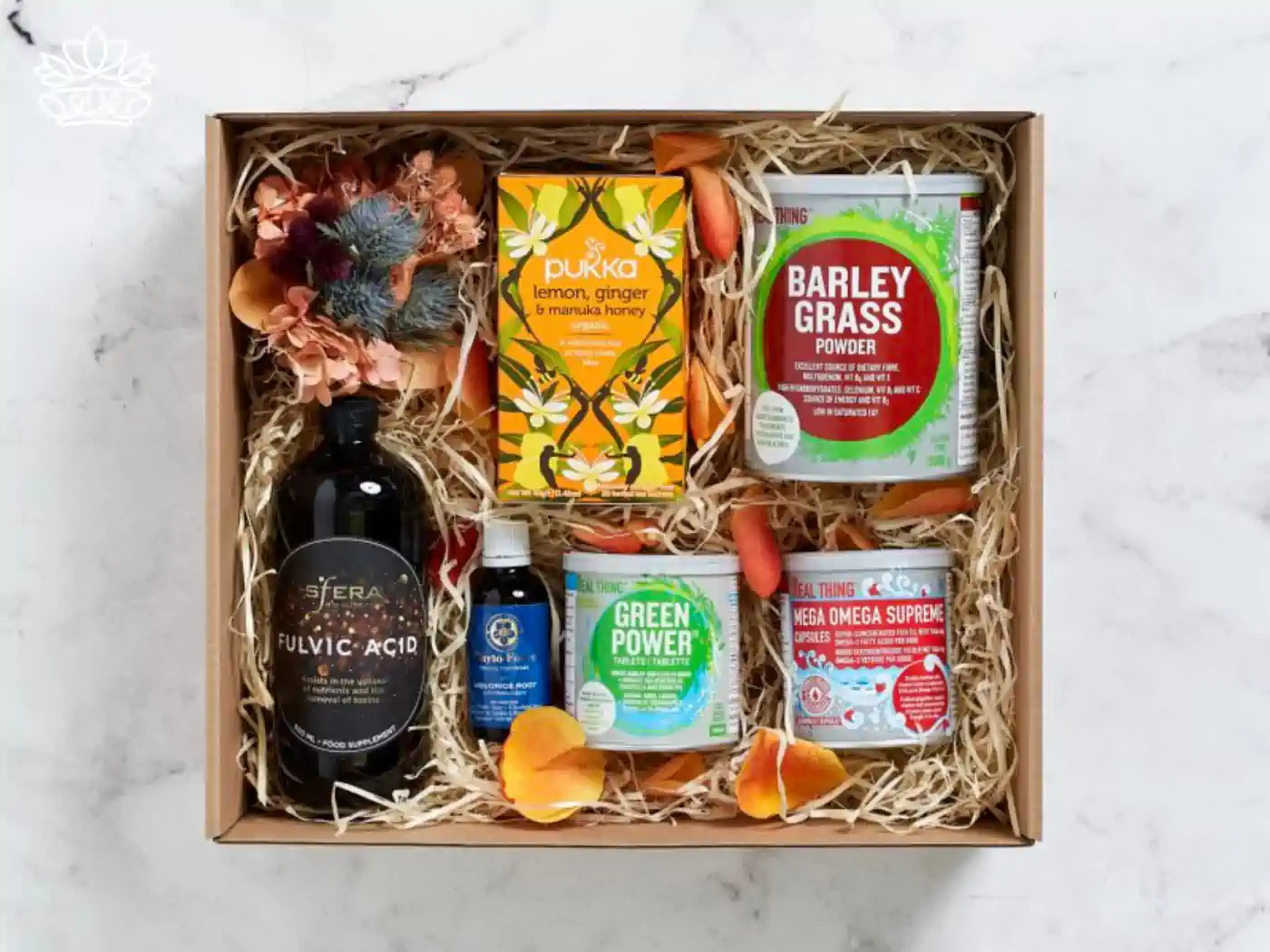 A curated selection of health products in a gift box including Pukka lemon, ginger & manuka honey tea, Barley Grass powder, Mega Omega Supreme capsules, Green Power blend, and Fulvic Acid supplement, surrounded by natural packing and decorative flowers, from the mental health gift boxes by Fabulous Flowers and Gifts. Carefully selected to boost wellness, delivered with deep care and consideration.