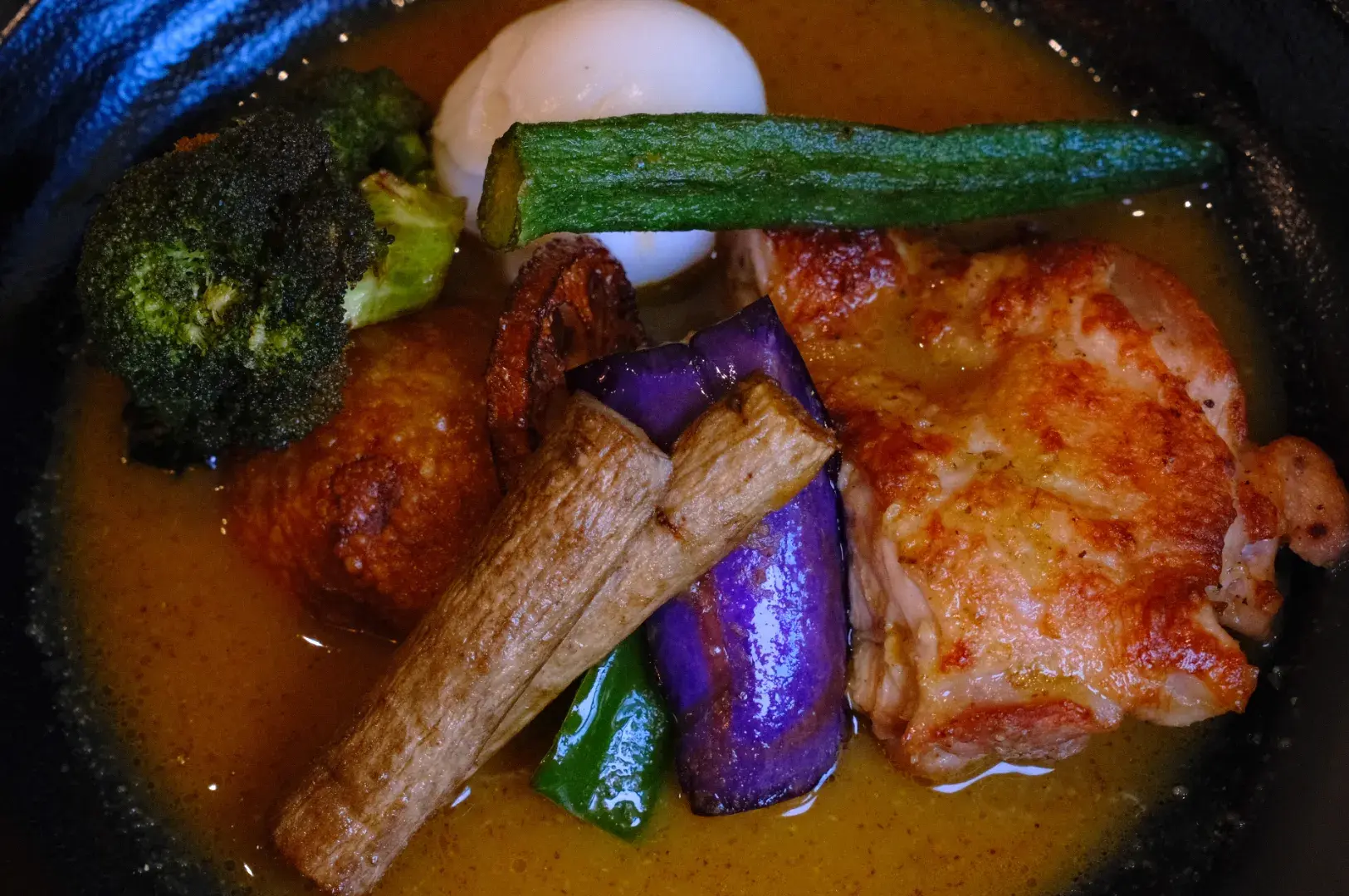 A bowl of orange curry with browned chicken, broccoli, zucchini, eggplant, mushrooms, and a boiled egg, arranged in a hearty, colorful meal.