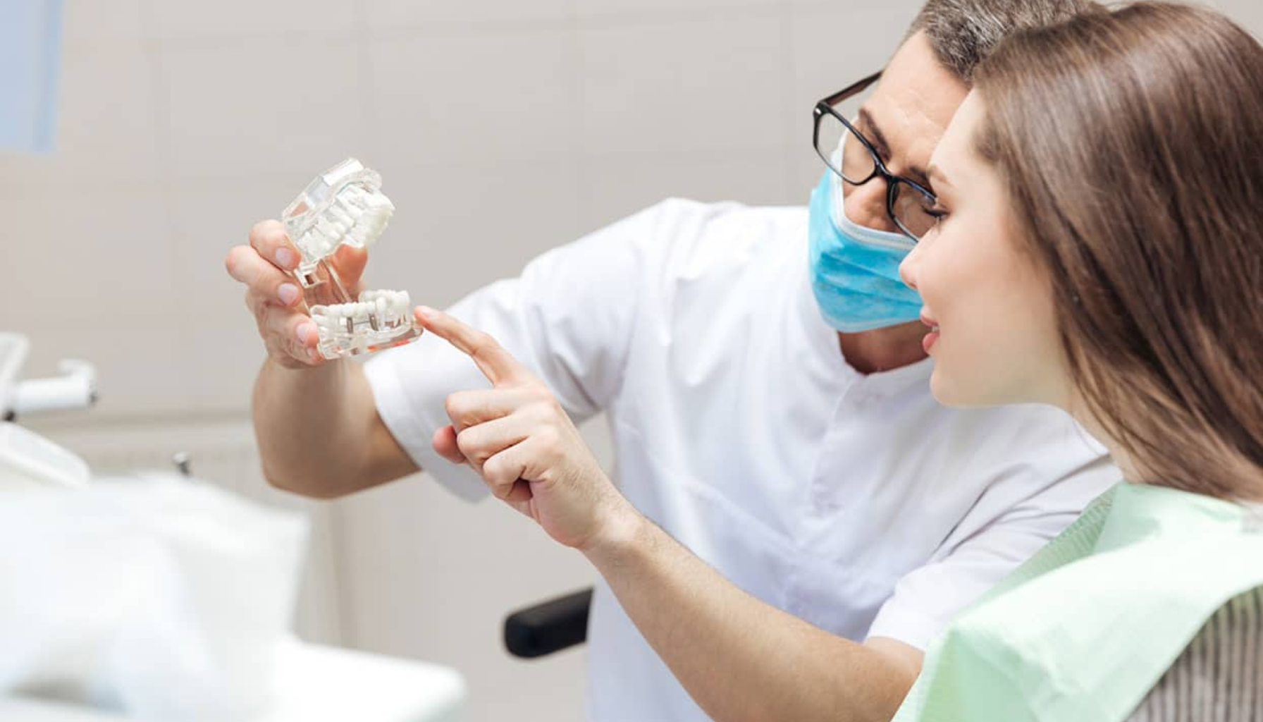 Tannlege forklarer tannbehandling ved hjelp av en tannmodell til en smilende pasient i tannlegestolen. tannklinikk tannklinikker tannbehandling våre offentlige telefon time andre