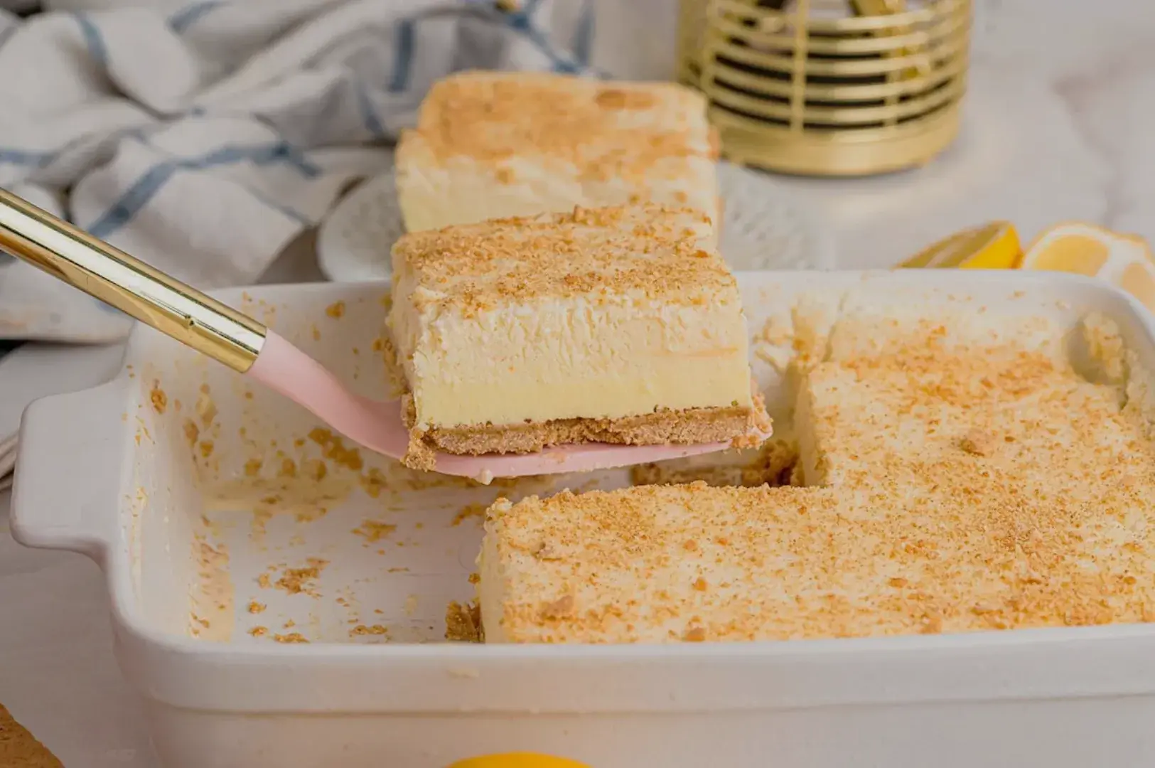 A close-up of a dessert with a graham cracker crust and creamy filling, with a slice being served from a white baking dish.