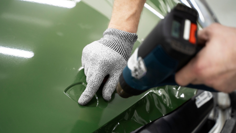 A mechanic is applying car wrap vinyl over rear bumper with the help of heat gun