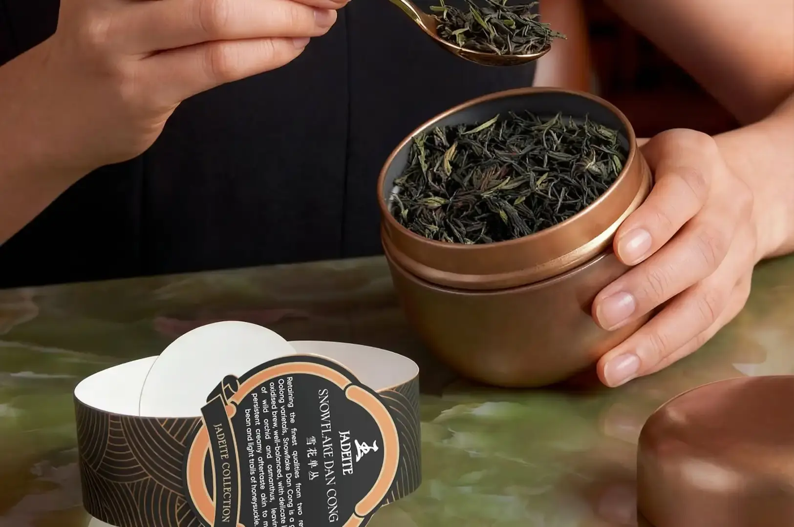 A close-up shot captures hands scooping dark green loose tea leaves with a brass spoon from an open, bronze-colored metallic tea caddy. The caddy sits on a green marble surface alongside its lid and a decorative paper band labeled "JADEITE SNOWFLAKE DAN CONG."
