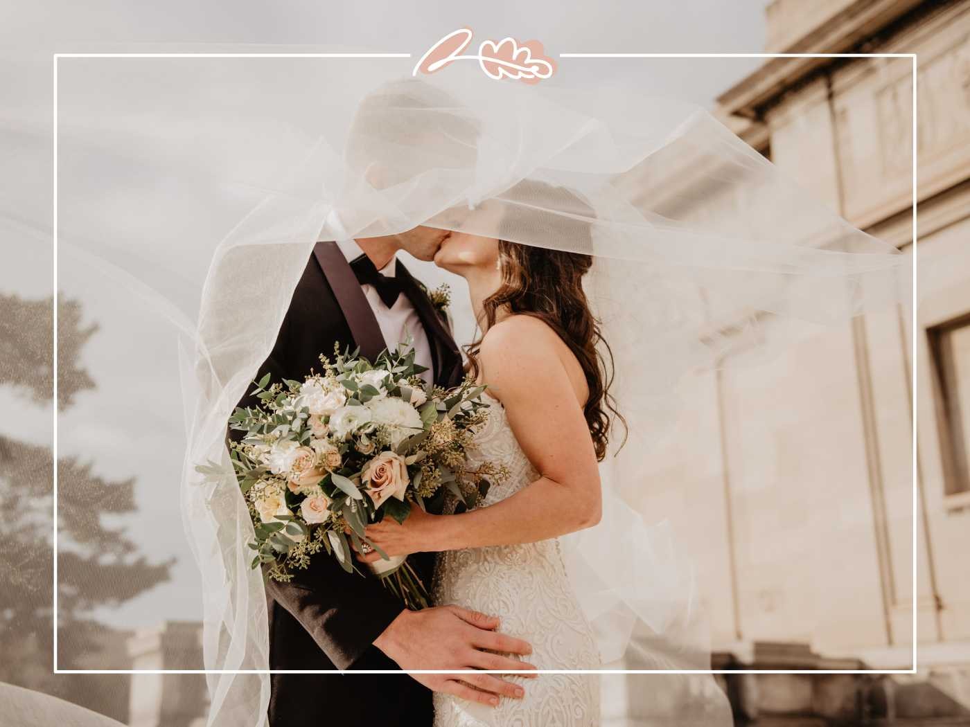 A bride and groom sharing a romantic kiss under the bride's veil, holding a beautiful bouquet of white and blush flowers. Fabulous Flowers and Gifts.